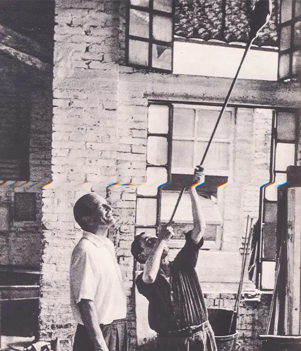 Black and white photograph showing two men (Paulo Venini) in an industrial or glass workshop setting, with one man reaching upward while holding a long pole or tool near a brick wall and large window panes.