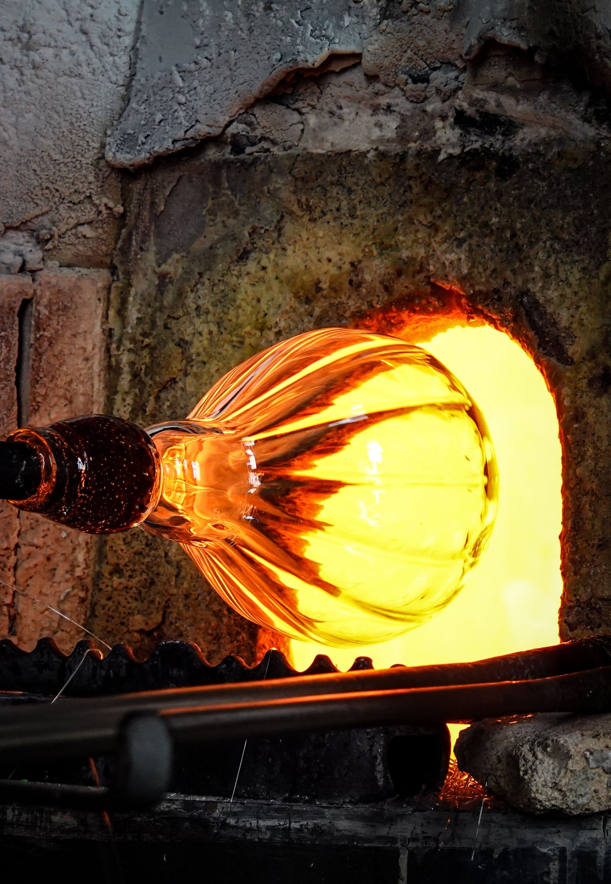 A glowing molten glass bulb being held by a blacksmith's tongs inside a traditional brick furnace, with bright orange and yellow light reflecting off the translucent glass.