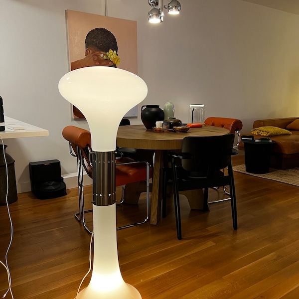 A vintage mushroom-shaped floor lamp b Carlo Nason with a frosted white top and base and a metallic silver central column, standing in a living room with wooden floors, a dining table, and chairs.
