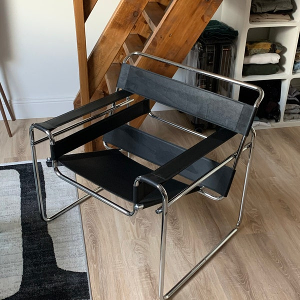 A metal and fabric chair "Wassily" with a chrome tubular frame and black woven seat and back straps, photographed in a real interior room with wood flooring, shelving, and a staircase visible.