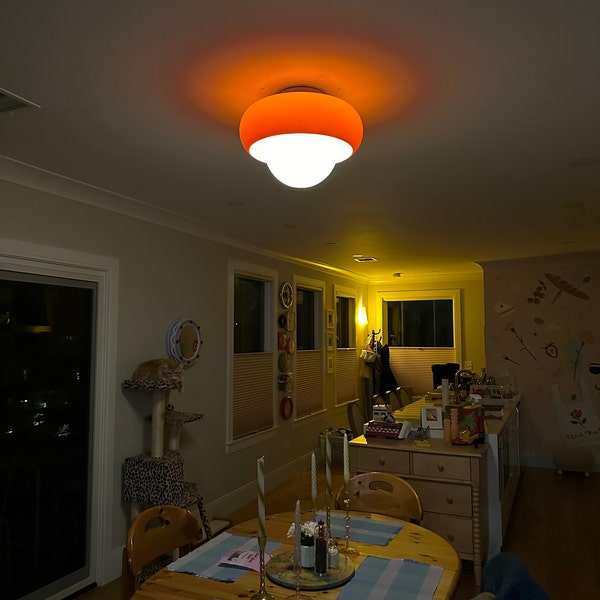 A vintage flush-mount ceiling light with a warm orange dome shade and a white frosted glass diffuser, glowing in a dining room setting.