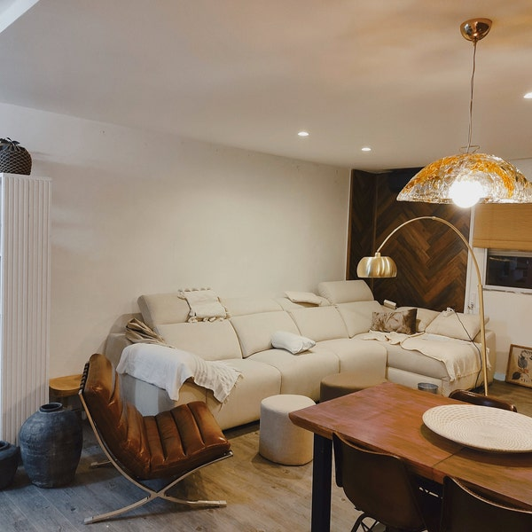 A vintage mid-century modern living room featuring a cream-colored sectional sofa, a brown leather lounge chair, a wooden dining table with chairs, and a large arched floor lamp with gold-toned shades.